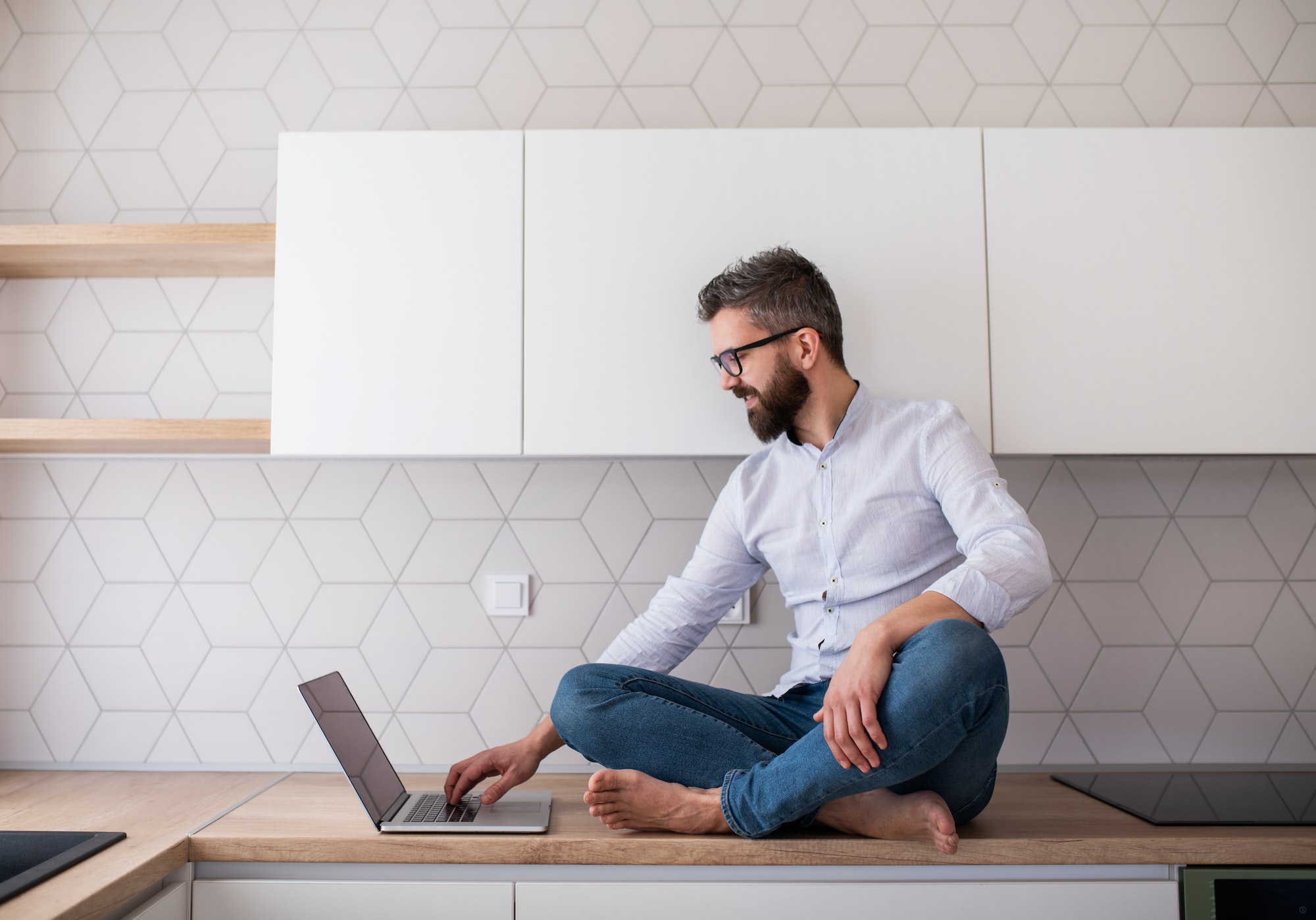 A mature man with laptop sitting in kitchen in unfurnished new house.