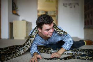 Down syndrome adult man sitting indoors in bedroom at home, playing with pet snake.