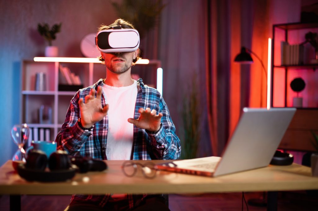 Young caucasian man sitting at home office and using VR glasses and laptop for remote work.